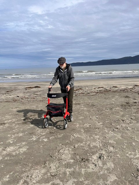 An older man using a walker on a sandy New Zealand beach under cloudy skies